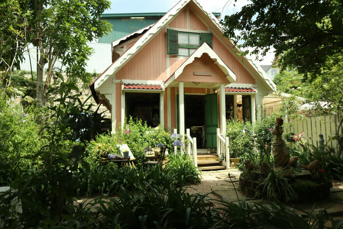 Pink wooden cottage with green shutters sits in a lush garden.